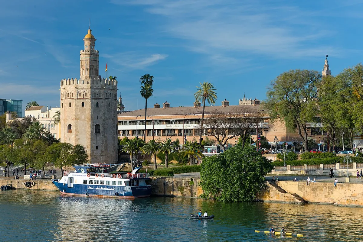 Torre del Oro beside the Guadalquivir in Seville, with a moored passenger boat and paddlers on the river.