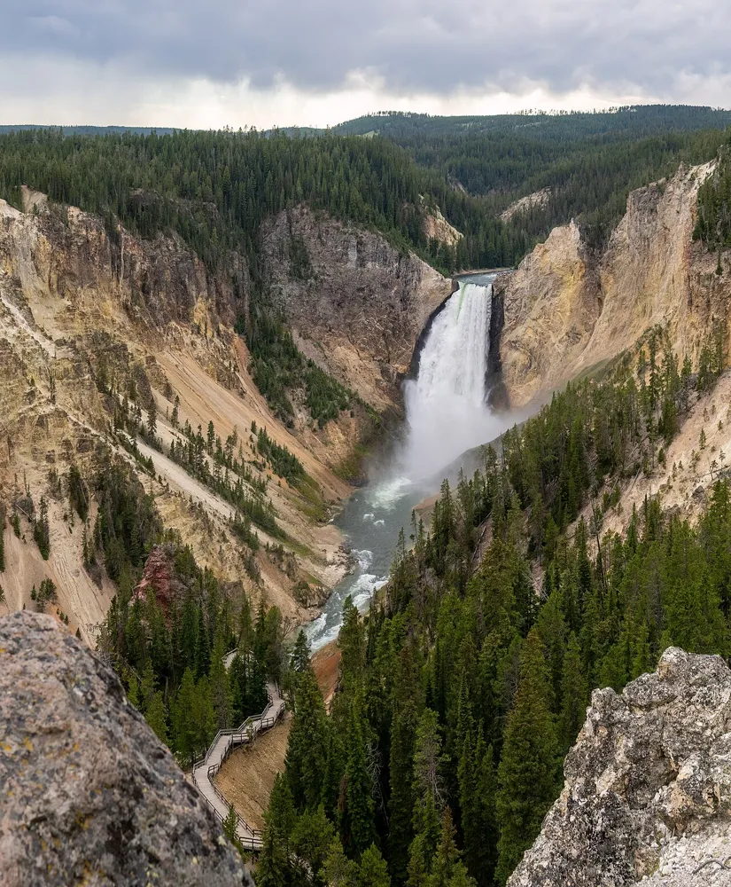 A waterfall plunges into a steep, forested canyon with pale yellow cliffs and a winding river below.