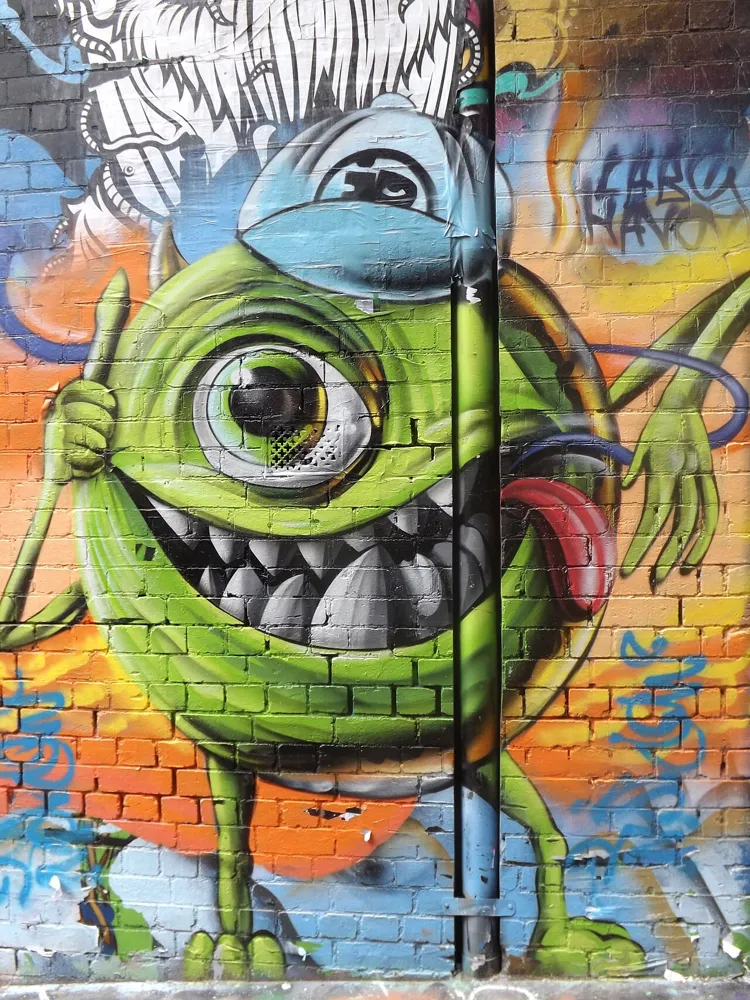 Street art of a grinning green, one-eyed character wearing a blue cap, painted on a brick wall.