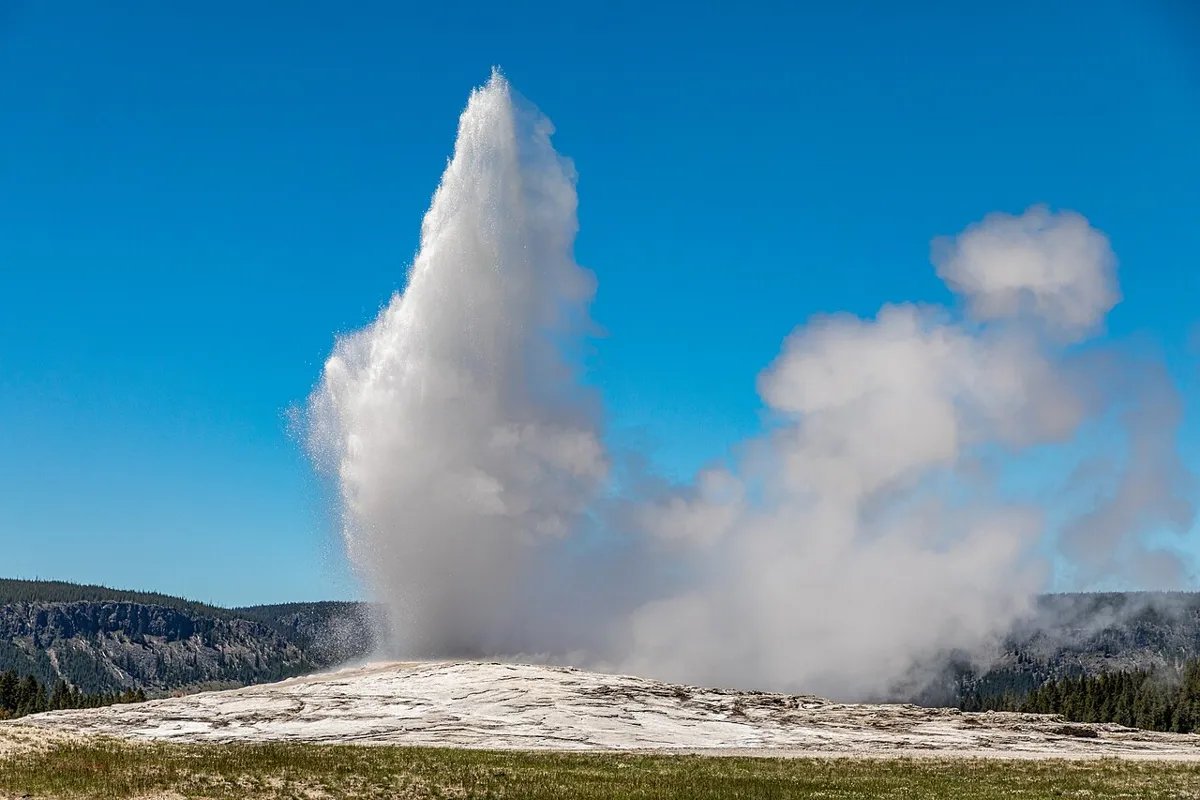 Old Faithful geyser erupting above a pale mineral mound, with steam drifting across a blue sky.