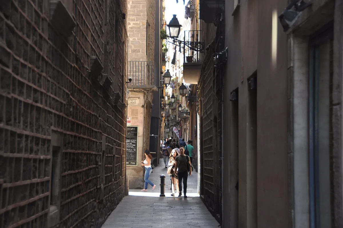 Pedestrians in a narrow stone-paved lane between historic buildings, with balconies and wall-mounted lanterns.