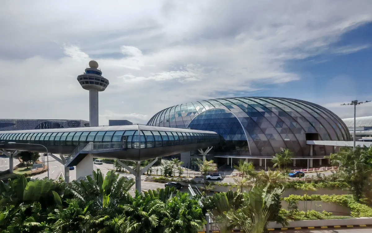 Airport control tower beside a glass dome, elevated enclosed walkway and tropical greenery.