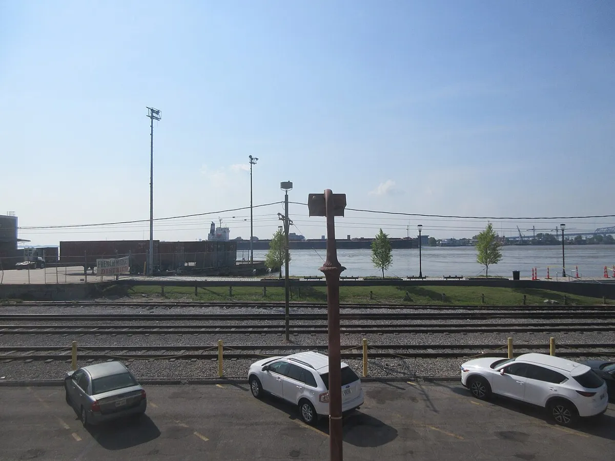 Parked cars and railway tracks in front of a grassy riverbank, with ships across the Mississippi River.