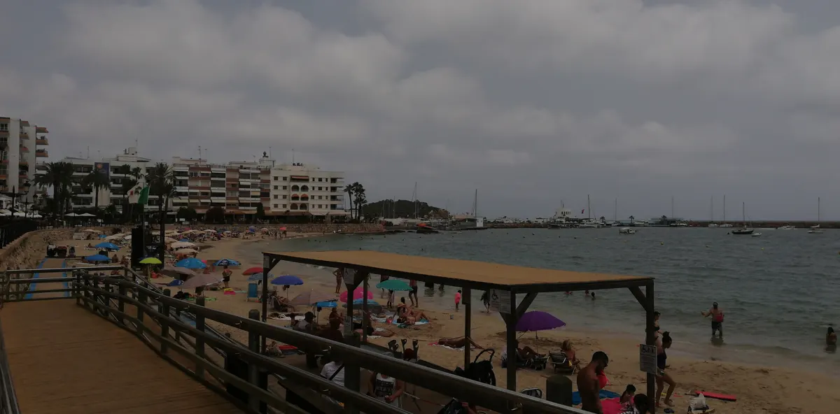 Beach with colourful umbrellas, swimmers, a wooden promenade and boats beneath cloudy skies.