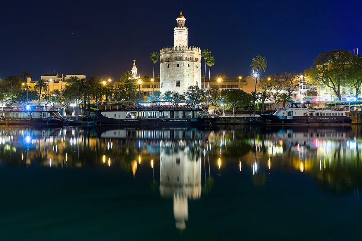 Illuminated Torre del Oro beside the Guadalquivir at night, with moored boats and reflections in the water.