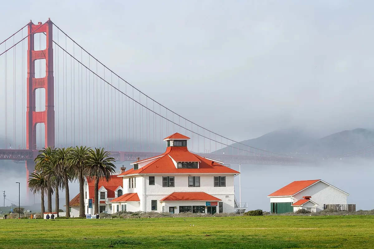 White buildings with red roofs and palm trees on a grassy waterfront, with the Golden Gate Bridge behind them in fog.