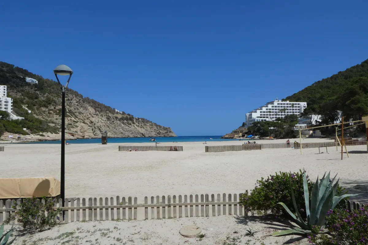 Wide sandy beach beside a blue bay, with rocky hills, white buildings and plants in the foreground.