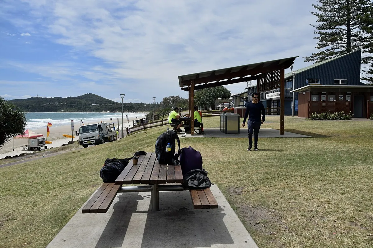 Picnic tables and a shaded shelter on a grassy beachfront beside Main Beach, with surf and a distant headland.