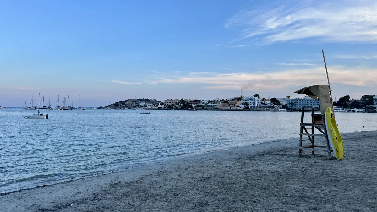 Sandy beach beside a calm bay with anchored sailboats, waterfront buildings and a lifeguard chair.