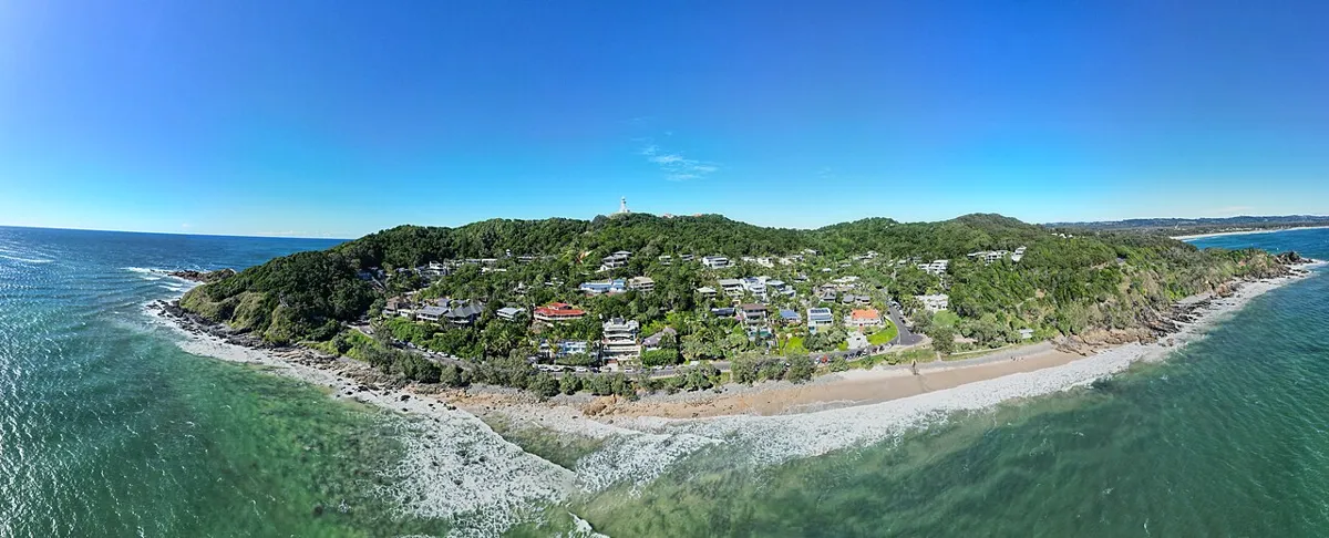 Panoramic view of Wategos Beach, with breaking waves, coastal homes and a wooded headland topped by a distant lighthouse.