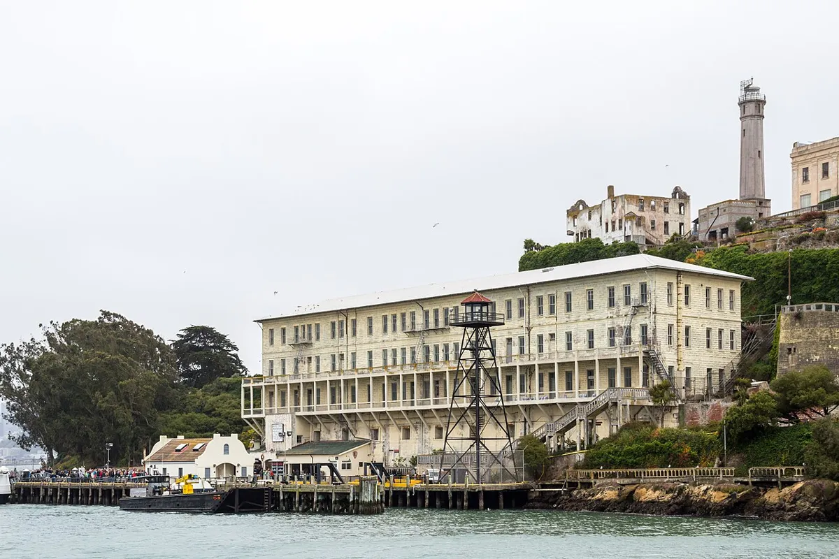 Alcatraz waterfront with a long pale building, a guard tower, a pier and a lighthouse on the hillside.