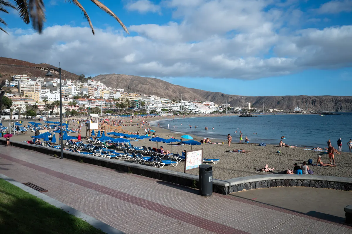 Beach with blue parasols, sun loungers, a paved promenade and buildings beneath dry hills.