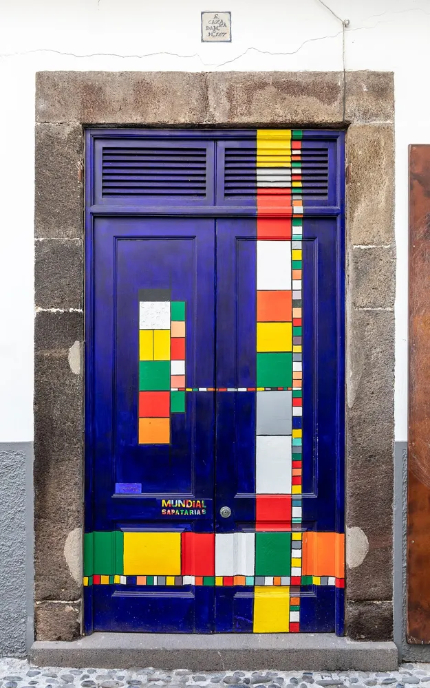 Deep blue double door painted with colourful geometric blocks, framed by stone with cobbled paving below.